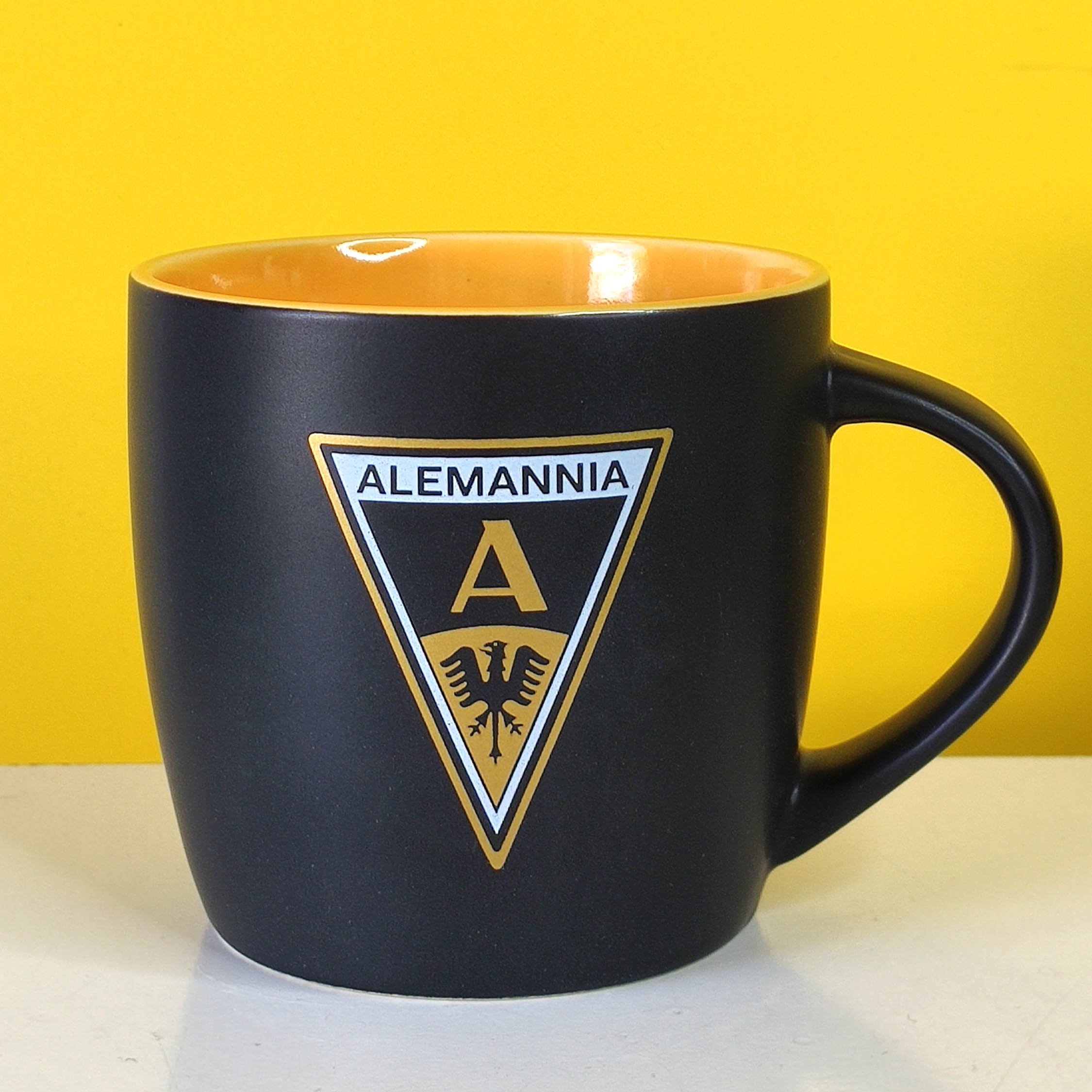 Alemannia cup - wappen Foto1 - Alemannia Shop Close-up of a black mug with a yellow interior and a shield-shaped emblem on the front, set against a bright yellow background.