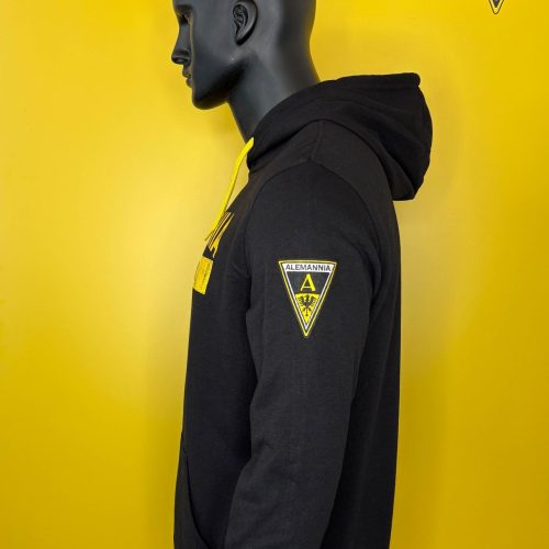 Side view of a black hoodie on a mannequin, with a yellow and black Alemannia patch on the sleeve and chest, against a yellow backdrop.