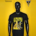 Mannequin wearing a black Alemannia graphic tee with bold yellow text and a soccer illustration, set against a bright yellow background; Alemannia logo in the top-right corner.