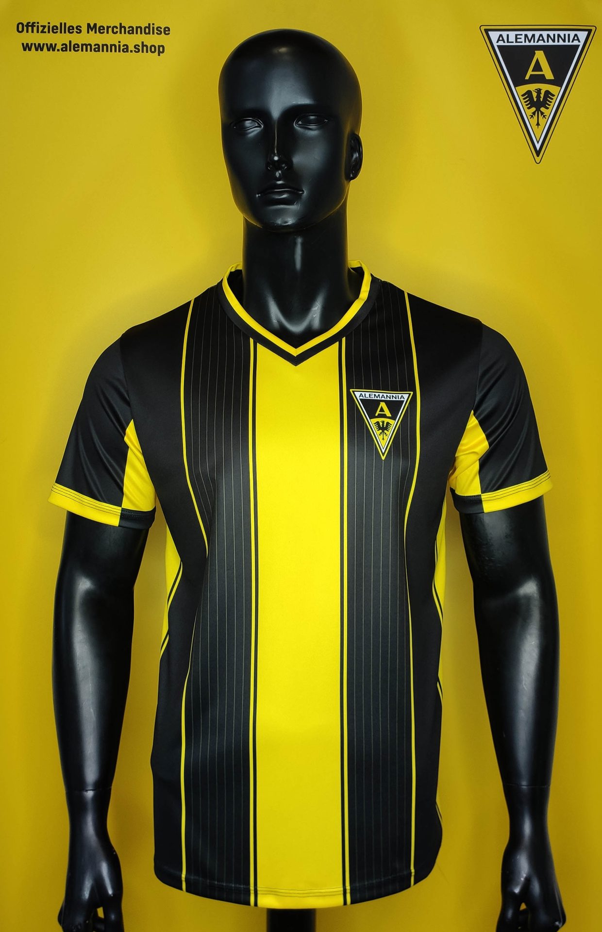 Alemannia Aachen - Stripe design shirt Foto1 - Alemannia Shop Black mannequin wearing a yellow and black striped soccer jersey with an Alemannia crest on the chest against a yellow background