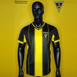 Black mannequin wearing a yellow-and-black striped soccer jersey with club crest on the chest against a yellow backdrop