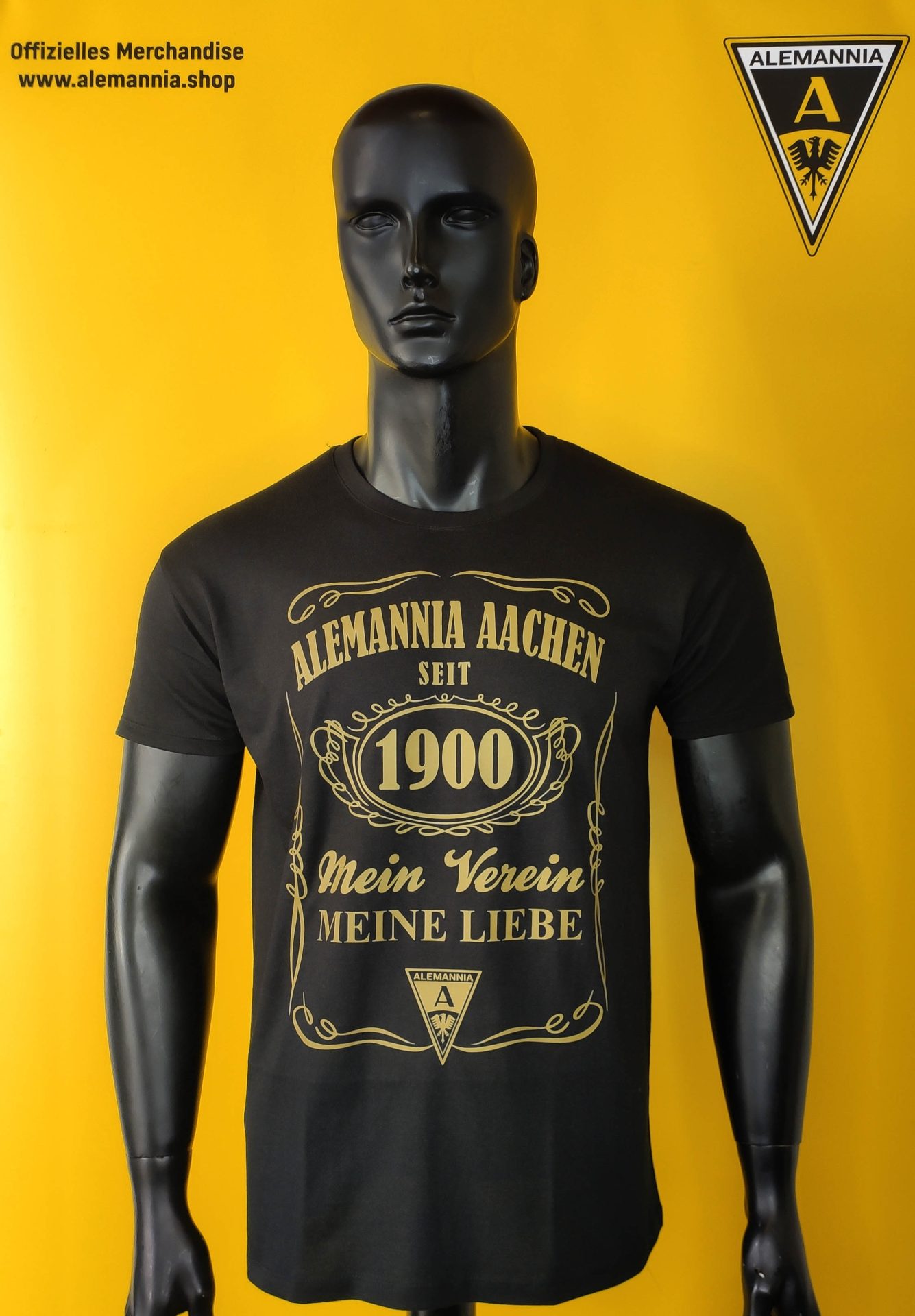 AATS63 Foto - Alemannia Shop Black T-shirt on a gray mannequin against a bright yellow background; shirt reads 'ALEMANNNIA ACHEN SEIT 1900 Mein Verein Meine Liebe' with emblem.