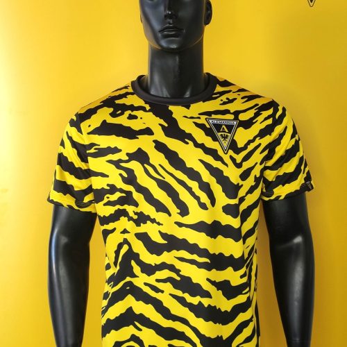 Mannequin wearing a yellow and black tiger-stripe jersey with an Alemannia crest on the chest against a yellow background.