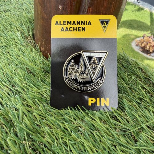 IMG_9295 - Alemannia Shop Close-up of an Alemannia Aachen pin on a black card, resting on grass beside a wooden post; shows the club crest and a cityscape design.