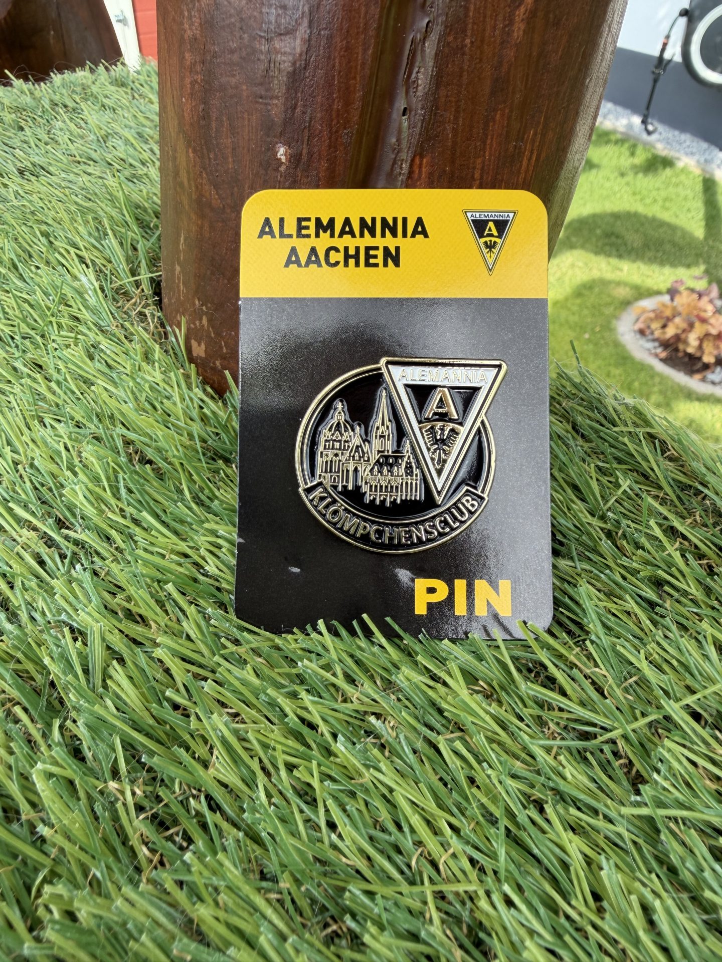 IMG_9295 - Alemannia Shop Close-up of an Alemannia Aachen pin on a black card, resting on grass beside a wooden post; shows the club crest and a cityscape design.