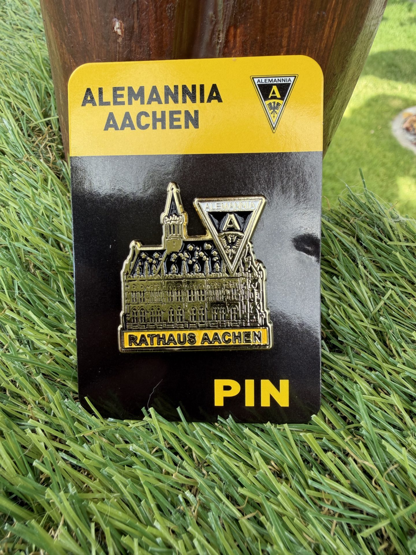 IMG_9296 - Alemannia Shop Gold-colored pin featuring Aachen's town hall illustration and Alemannia Aachen crest, attached to a black and yellow backing plate (RATHAUS AACHEN)