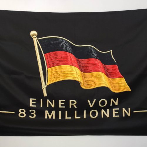Black banner with a gold-embroidered German flag waving, held at the corners by hands, showing the text 'EINER VON 83 MILLIONEN'