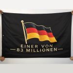 Black banner being held at the corners, featuring an embroidered German flag and the text 'EINER VON 83 MILLIONEN'.